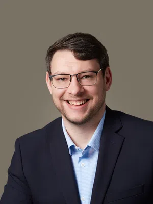 Smiling man with glasses wearing a navy blazer and light blue shirt against a beige background