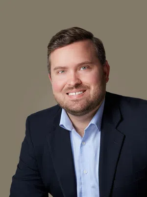 Smiling man with short brown hair and beard wearing a navy blazer and light blue button-up shirt against plain background