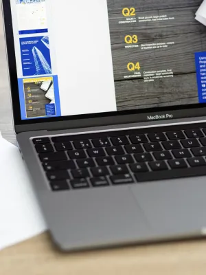 MacBook Pro displaying project management timeline with architectural plans on table.
