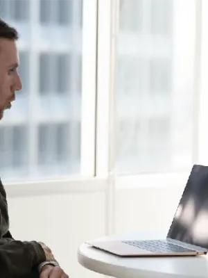 Man in wheelchair working on laptop at round table near large windows in bright office space
