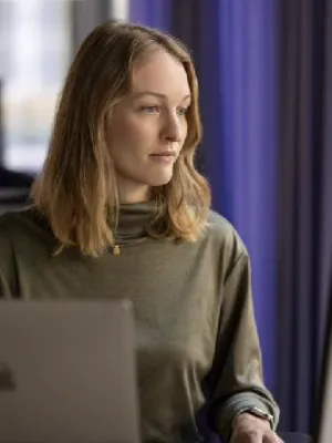 Woman in green sweater working on a laptop and desktop computer in a modern office with purple curtains.