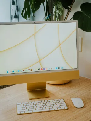 Yellow iMac computer on wooden desk with wireless keyboard and mouse, green plant in background.