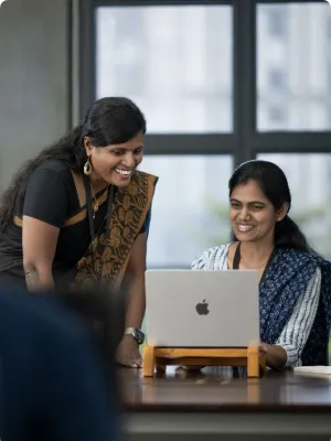 Two women smiling and collaborating over a laptop in a modern office setting by a large window.