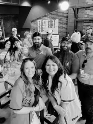 Group of smiling people at a social gathering indoors, some wearing sports jerseys, holding drinks, and posing for a photo