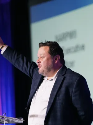 Man in suit speaking at podium with raised hand during presentation against purple and white background