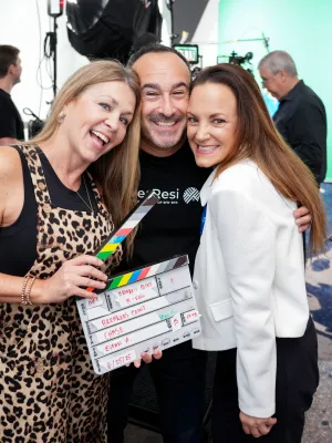 Three people smiling and holding a clapperboard on a film set with studio lights and green screen background
