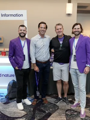 Four men smiling at an event, with two wearing purple jackets in front of an information desk for AppFolio and Second Nature.