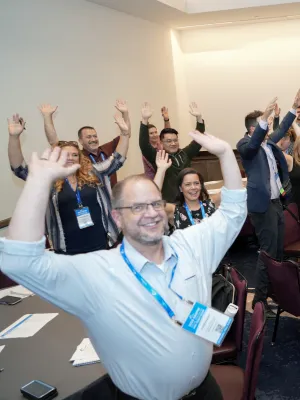 Group of conference attendees smiling and raising hands around tables in a meeting room with lanyards and badges.