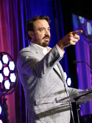 Man in gray suit speaking and pointing on stage with purple curtains and large screen backdrop.