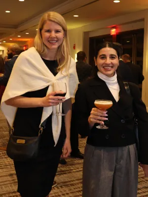 Two women smiling and holding drinks at an indoor social event with warm lighting and carpeted floor