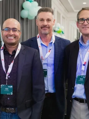 Three men wearing business casual attire and conference badges smiling at an indoor event with balloons in the background.