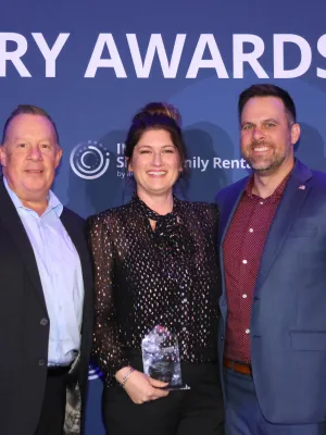 Three smiling people at Industry Awards event, one holding a clear glass trophy against blue backdrop.
