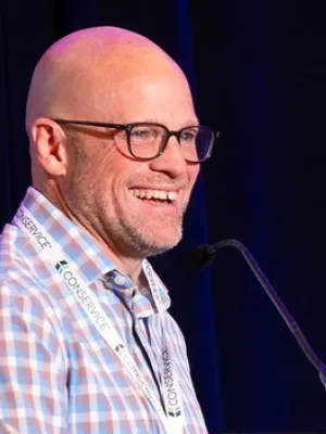 Smiling bald man wearing glasses and checkered shirt speaking at a microphone on stage with dark blue background