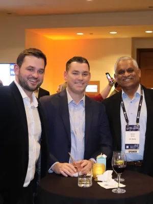 Four men in business attire smiling and posing around a cocktail table at a conference event indoors.