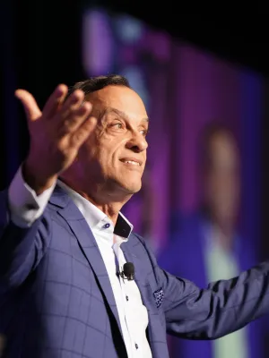 Man in blue suit speaking on stage with arms outstretched and purple-blue light background