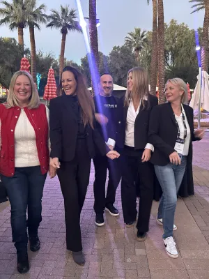 Group of five people smiling and walking outdoors near a pool with palm trees and colorful umbrellas at dusk.