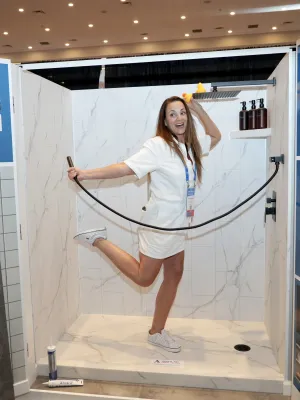 Woman demonstrating Palisade waterproof grout-free shower wall system in a marble-style shower booth at a trade show.