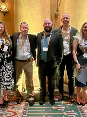 Group of seven professionals wearing conference badges posing in a warmly lit room with decorative carpet and wall lighting.
