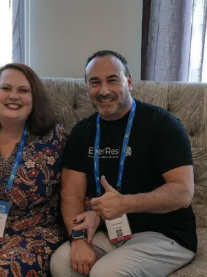 Two smiling conference attendees sitting on a sofa showing thumbs up with event badges and blue lanyards