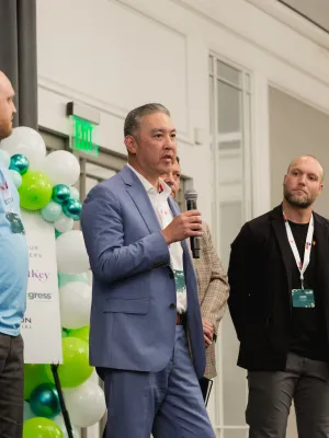 Group of five men in business attire speaking at an indoor event with balloons and signage in the background