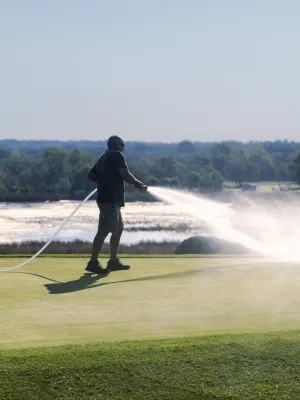 Person watering grass on a golf course with hose under sunny sky and natural landscape in the background