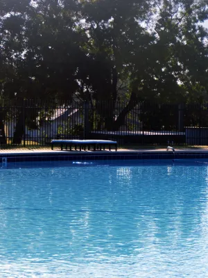Outdoor swimming pool with clear blue water surrounded by trees and a black metal fence on a sunny day.