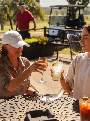 Two women enjoying drinks and toasting at an outdoor patio table on a sunny day at a golf course.