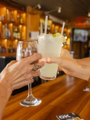 Two women clinking glasses with cocktails at a wooden bar in a cozy pub setting.