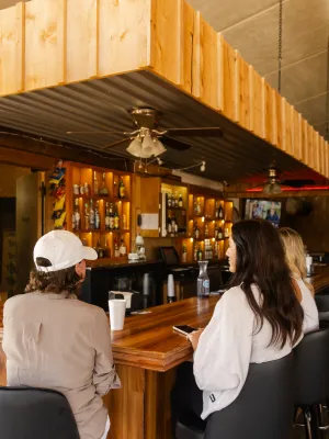 Three people sitting at a wooden bar counter in a cozy, warmly lit bar watching multiple wall-mounted TVs.