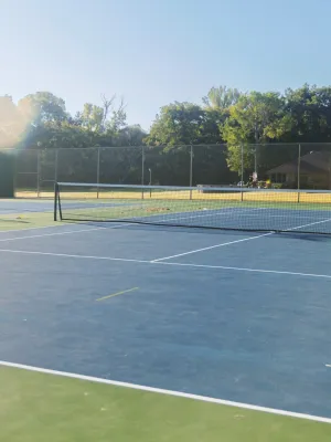 Empty outdoor tennis courts in sunlight with trees and buildings in the background on a clear day.
