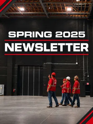 Four construction workers in red safety gear walk inside a large industrial warehouse with Spring 2025 Newsletter text.