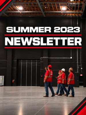 Four workers in red safety gear walking inside a large industrial warehouse under Summer 2023 Newsletter text.