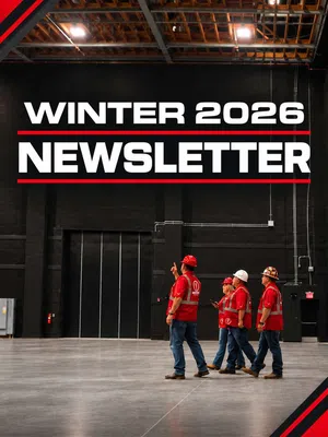 Four workers in red safety vests and helmets walk inside a large industrial warehouse under a Winter 2026 Newsletter sign.