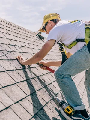 Roofer with safety harness installing shingles on a steep residential roof using a hammer and drill.