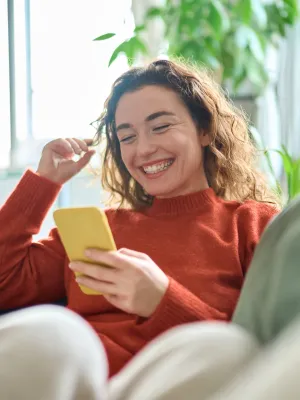 Smiling woman in orange sweater sitting on couch using yellow smartphone in bright living room with plants.