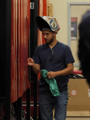 Man wearing welding helmet inspects large red industrial tanks in a workshop or factory setting.