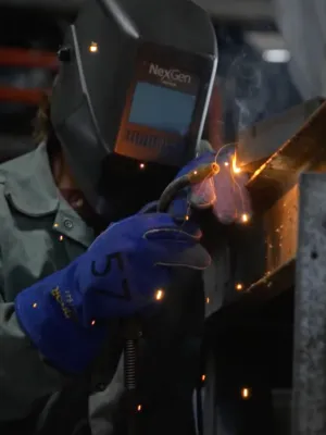 Welder wearing protective gear sparks flying while welding metal beam in industrial workshop setting.