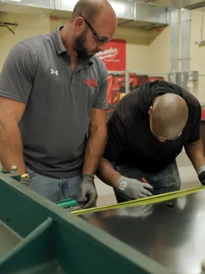 Two men measuring a large metallic sheet with a tape measure in an industrial workshop setting.