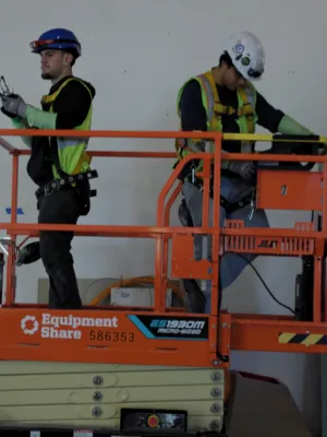 Two construction workers on an orange scissor lift inside a building under renovation with safety gear.