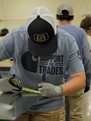 Young man wearing gloves measures and marks metal sheet in a workshop with others working in background.