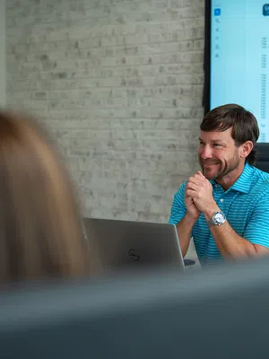 Man in blue striped shirt smiles during a meeting with colleagues in a modern office setting.