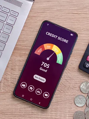 Smartphone displaying a 705 good credit score app, surrounded by laptop, calculator, and coins on wooden desk.
