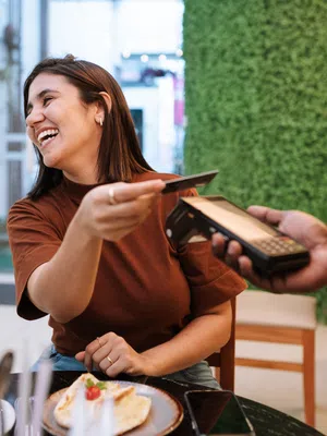 Smiling woman paying with credit card at cafe table with dessert and drink, card reader held by server.