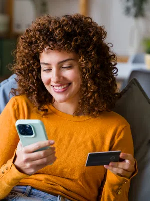 Smiling woman with curly hair using smartphone and credit card for online shopping at home.