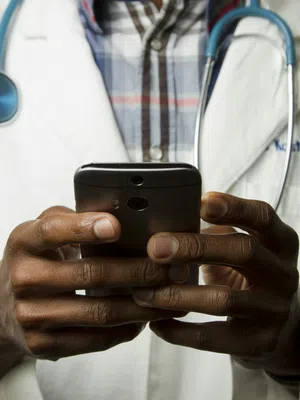 A doctor in a white coat checks a smartphone while wearing a stethoscope around their neck.