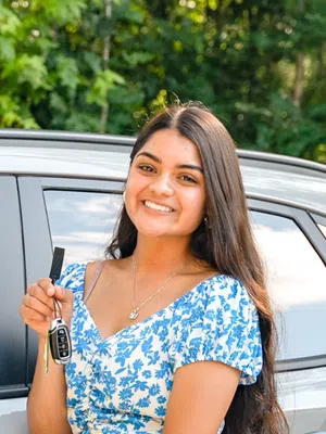 Smiling young woman holding car keys while standing next to a silver SUV with trees in background