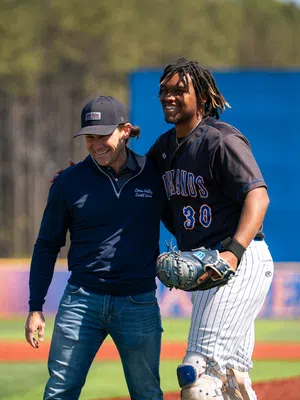 Two men smiling and embracing on a sunny baseball field with one in uniform and the other in casual wear.