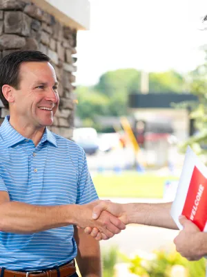 Two men shaking hands outside a building, one holding a welcome brochure, smiling and greeting.