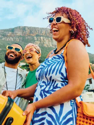 Group of friends laughing and packing an orange suitcase into a car with mountains in the background on a sunny day.
