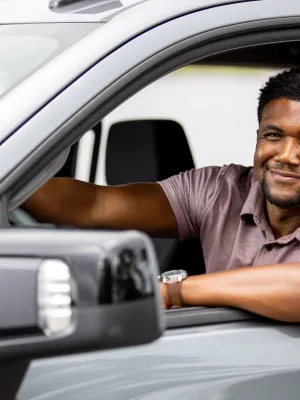 Smiling man in a purple shirt sitting in the driver's seat of a gray car with his arm resting on the window.